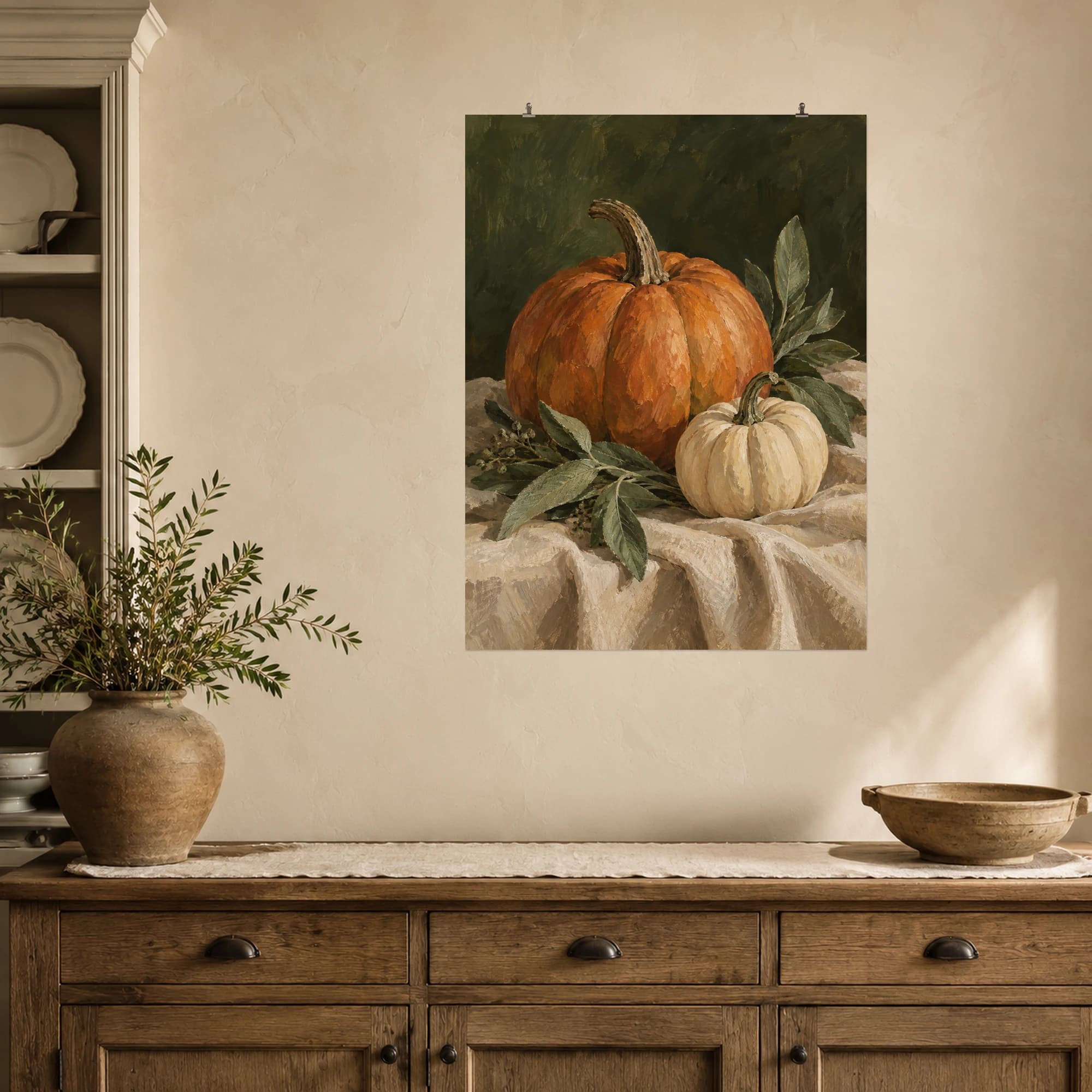 An orange pumpkin and a small cream pumpkin sit among sage leaves on warm linen. A dark olive backdrop brings depth to this calm autumn still life.