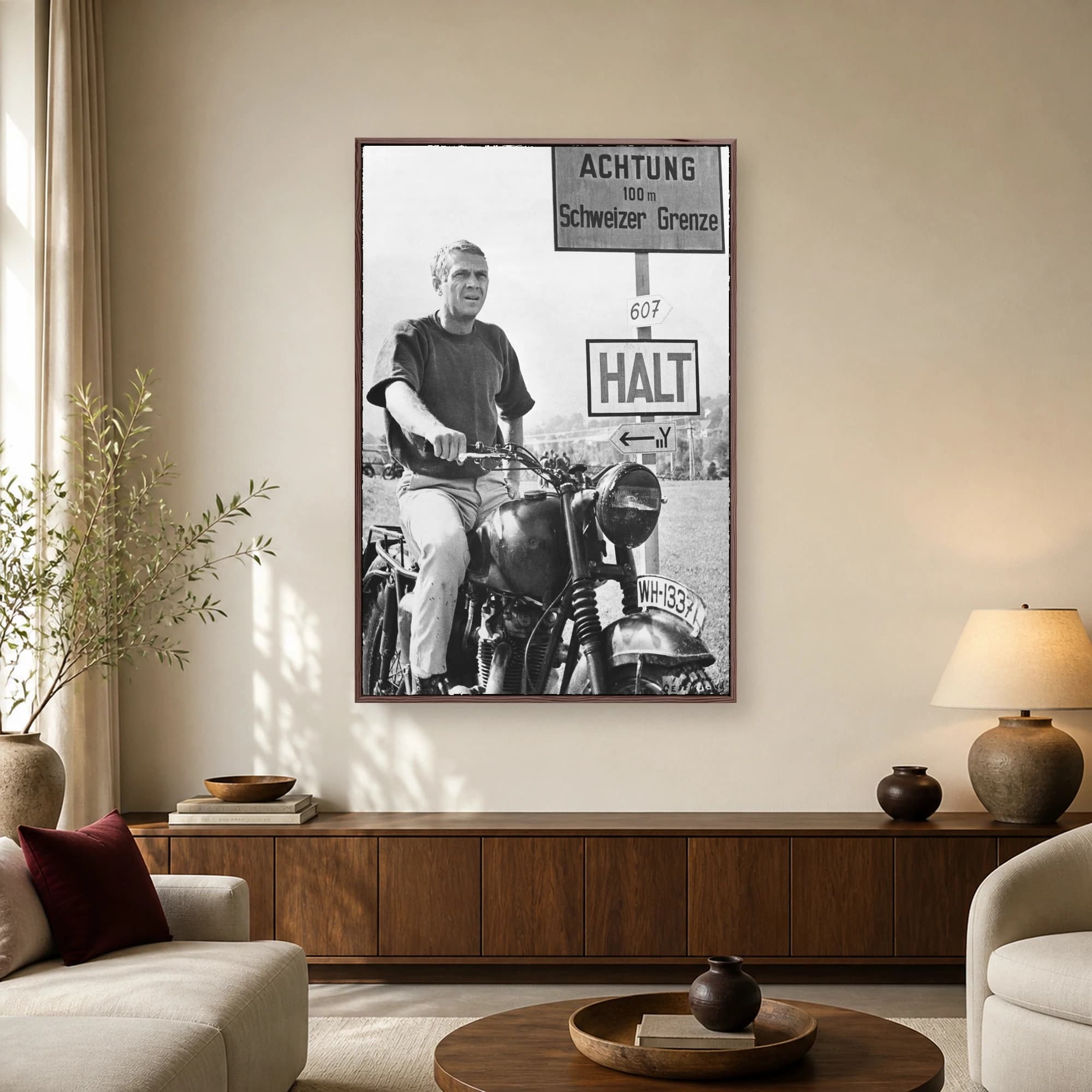 Black and white photograph of actor Steve McQueen sitting on a vintage motorcycle next to German border signs in a classic film scene.