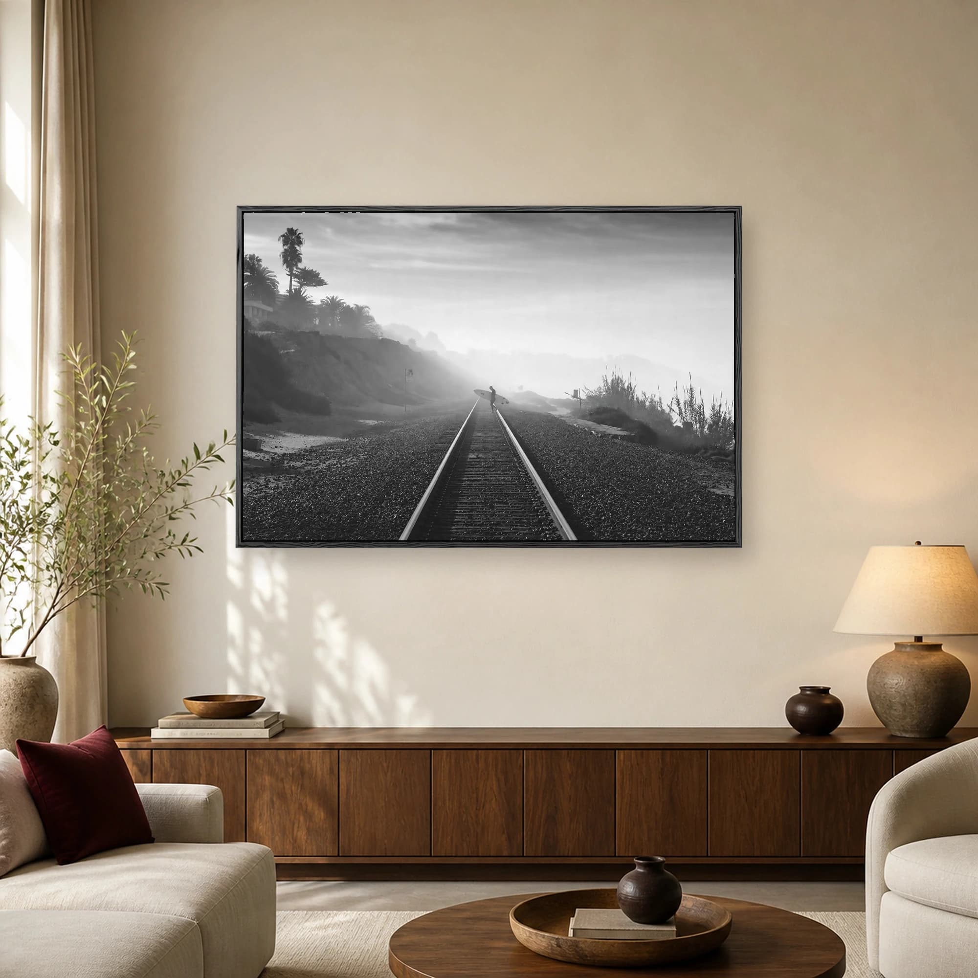 A black and white photograph of a lone surfer carrying a surfboard on railway tracks flanked by coastal cliffs and dense morning fog.
