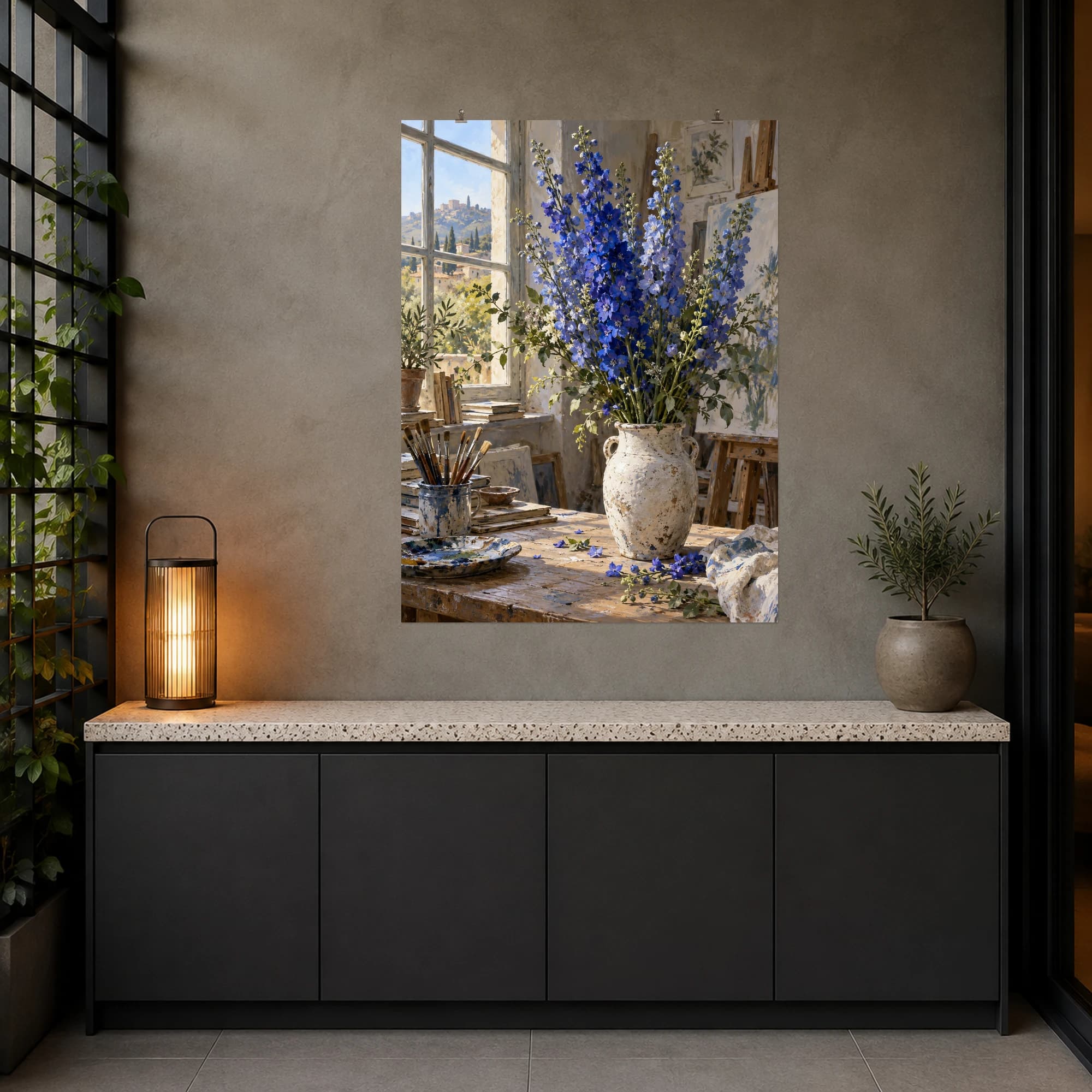 Blue delphiniums stand in a white vase in a softly lit studio. The tall flowers and worn work surface create an informal floral still life.