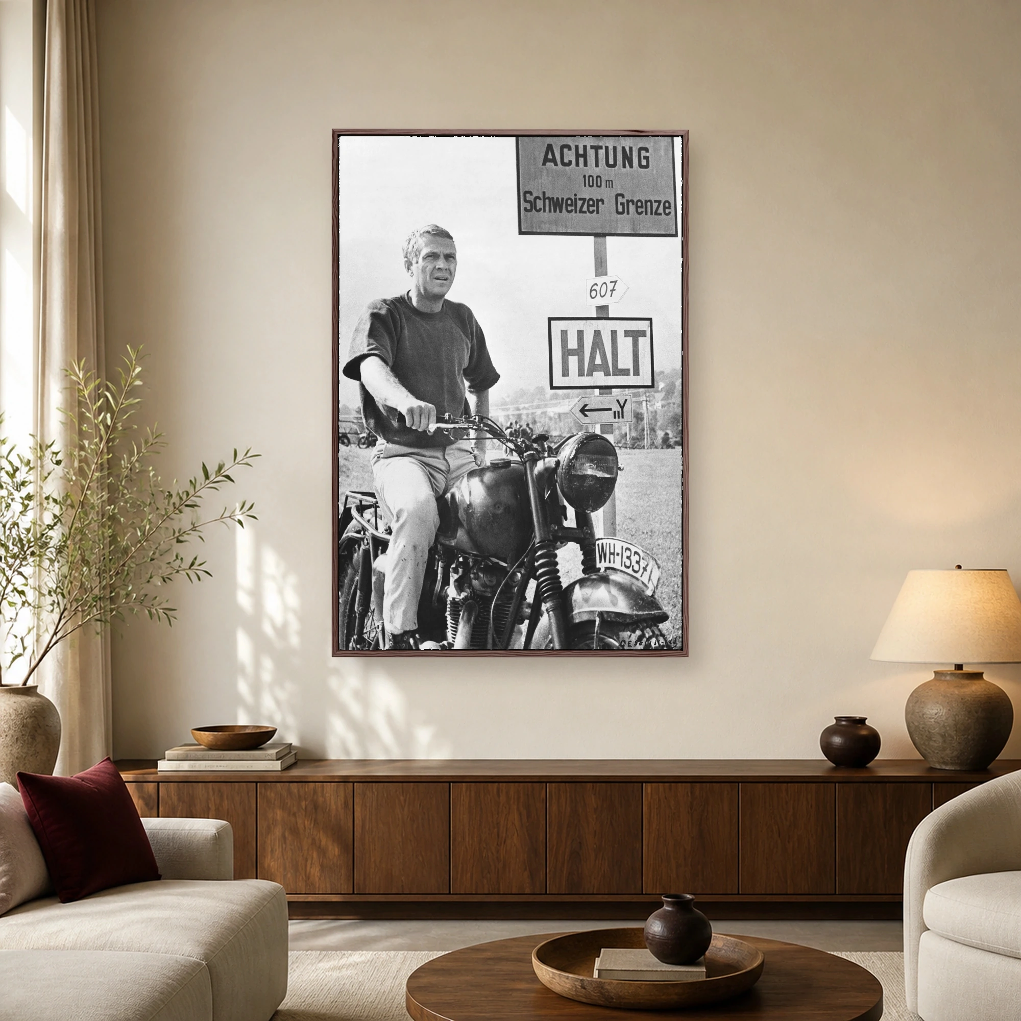 Black and white photograph of actor Steve McQueen sitting on a vintage motorcycle next to German border signs in a classic film scene.