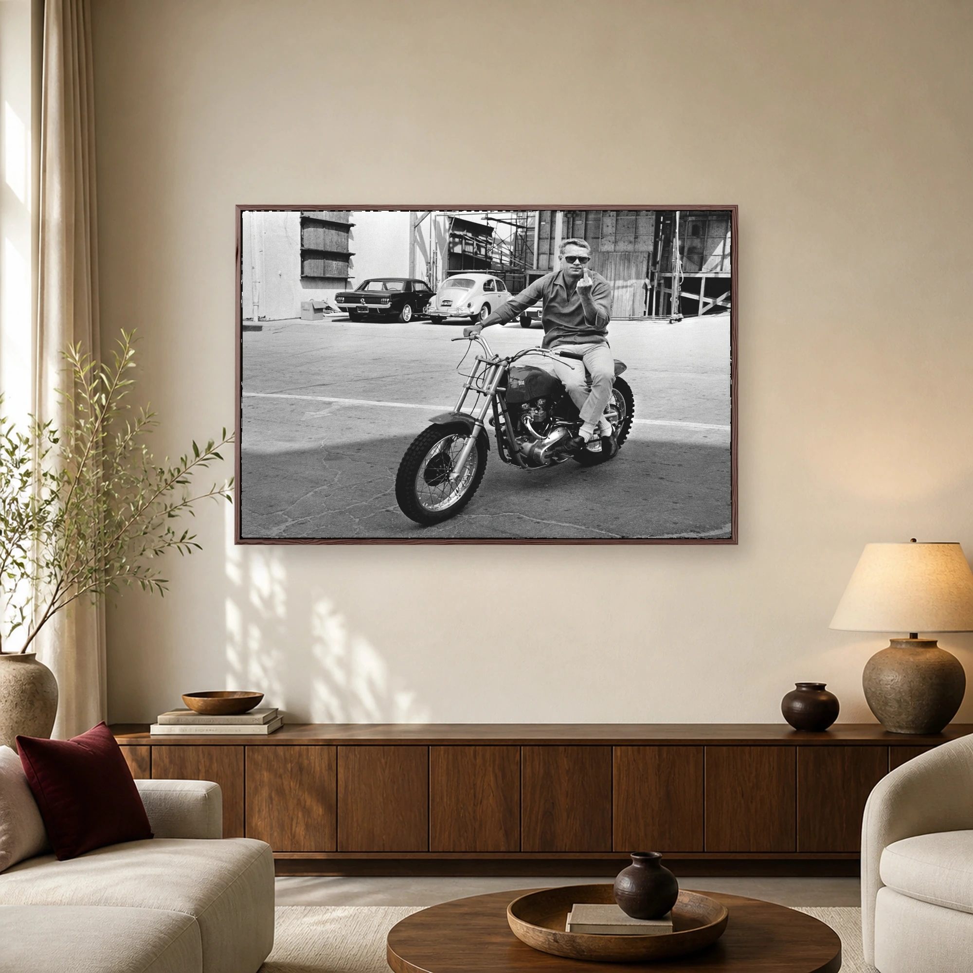 Black and white photograph of Steve McQueen wearing sunglasses on a vintage motorcycle making a defiant gesture on a studio lot.