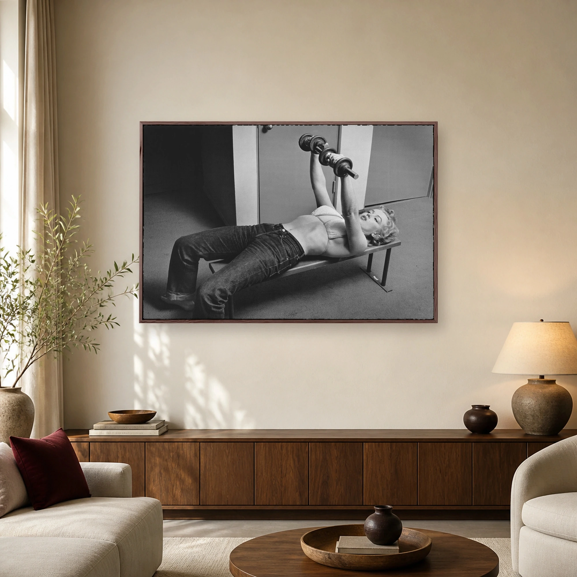 Black and white vintage photograph of Marilyn Monroe lying on a workout bench lifting a bar with dumbbells in high-waisted jeans and a terrycloth top.